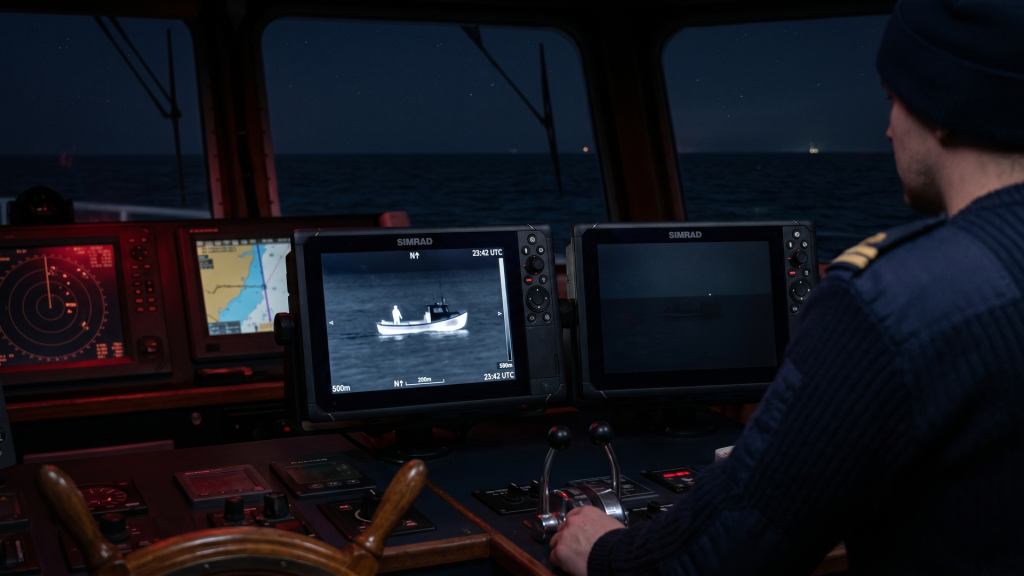 RMD-19-P image by Seatronx Marine vessel bridge at night with two rugged side-by-side monitors on the helm console. The left monitor shows a thermal imaging feed of a small unlit fishing skiff glowing as a warm shape against a cold black ocean; the right monitor shows the same view in visible light with the skiff almost invisible. A red-dimmed chart and radar display sit to the left of the watch officer, who stands at the wheel in low red night lighting.