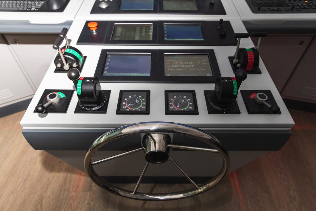 Crane Camera Systems image by Seatronx Modern commercial ship bridge console with a stainless steel steering wheel, throttle controls, instrument gauges, and a row of flush-mount marine navigation displays at the helm.