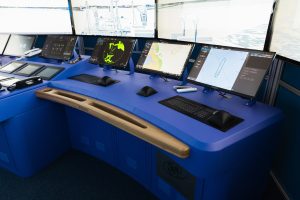 Modern commercial ship bridge with a blue navigation console showing multiple monitors with radar sweeps and electronic chart displays, with keyboards and helm controls visible.