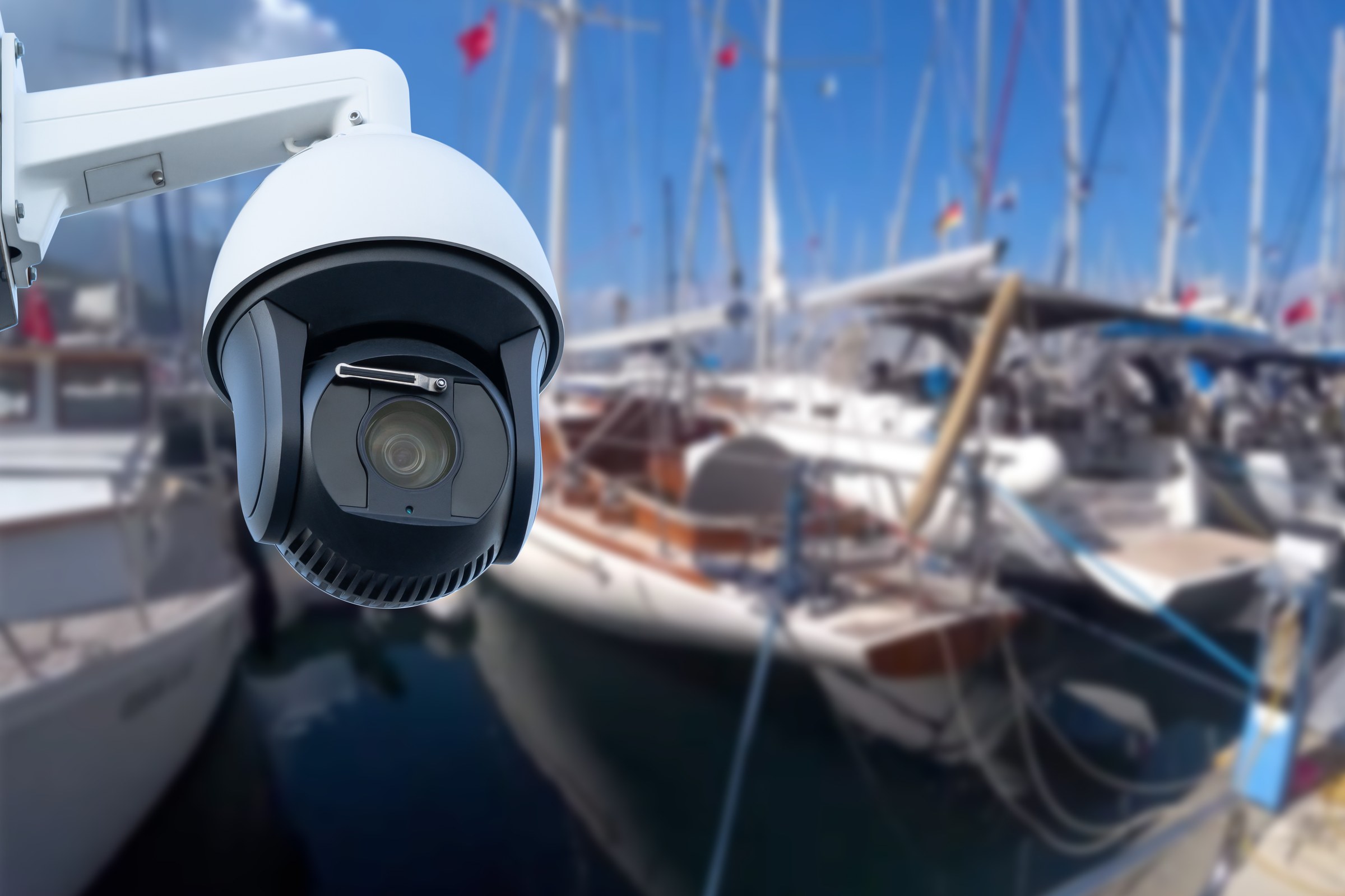 White pan-tilt-zoom CCTV dome camera on a wall bracket overlooking moored yachts in a marina