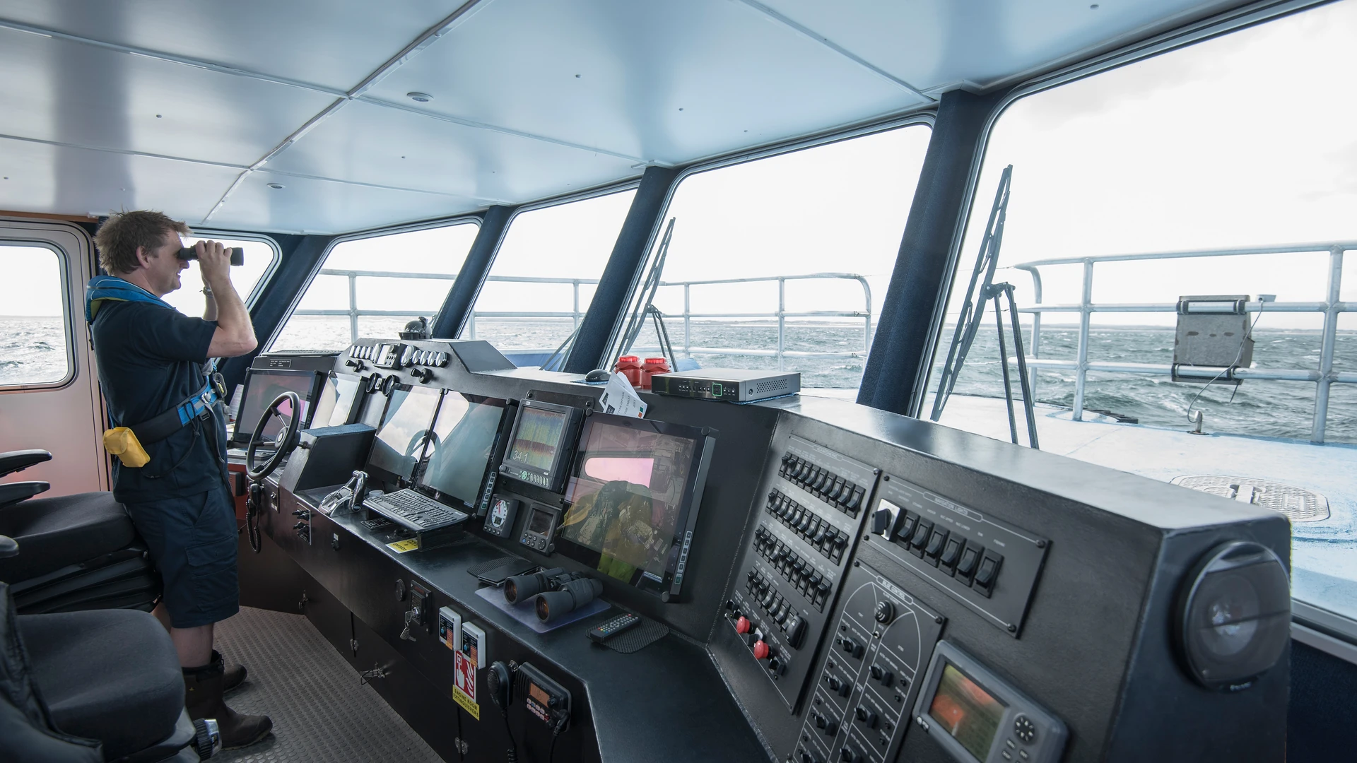 Person looking through binoculars on a vessel bridge lined with displays and switch panels