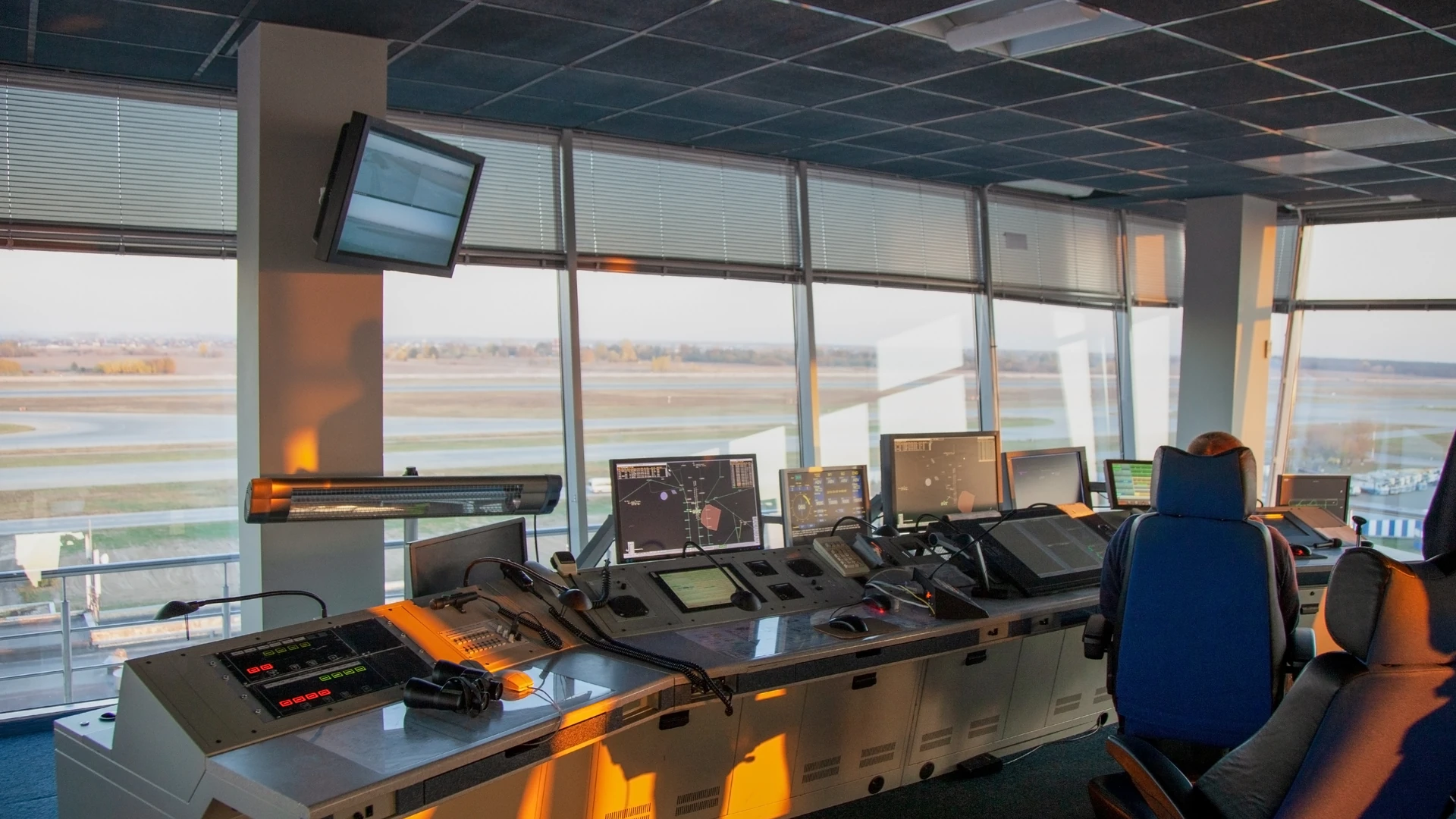 Tower cab with a console of radar screens, keypads, gooseneck microphones and a mouse, a person seated at the right and the airfield outside.