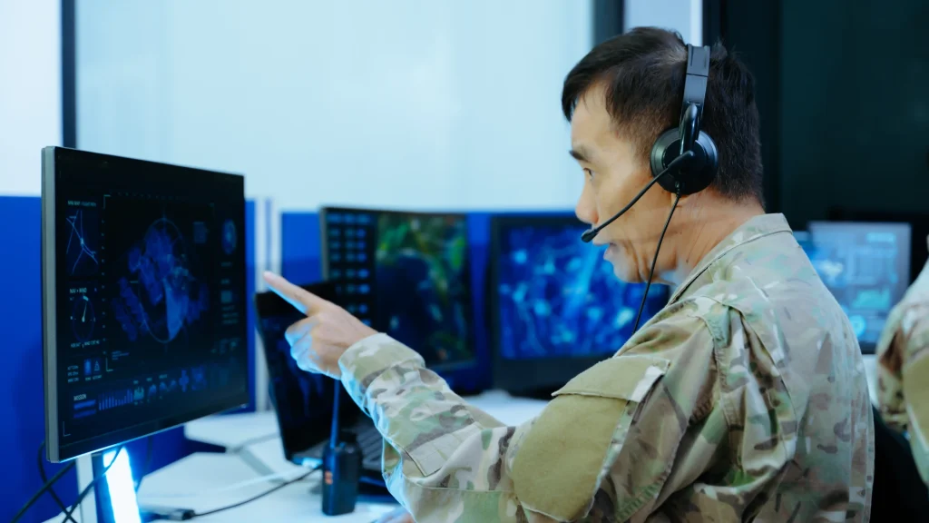 Markets image by Seatronx A person in a camouflage uniform and headset points at a monitor in a room with several screens, a laptop and a handheld radio on the desk.