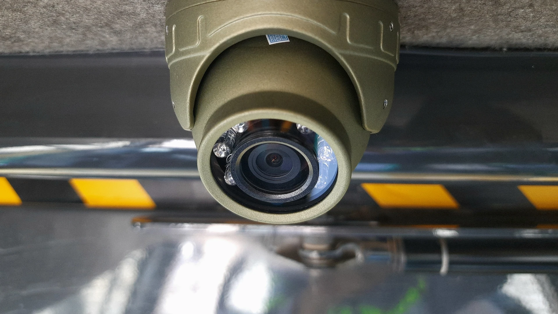 Close-up of an olive dome camera with infrared LEDs mounted on a vehicle cab ceiling above a yellow and black hazard stripe