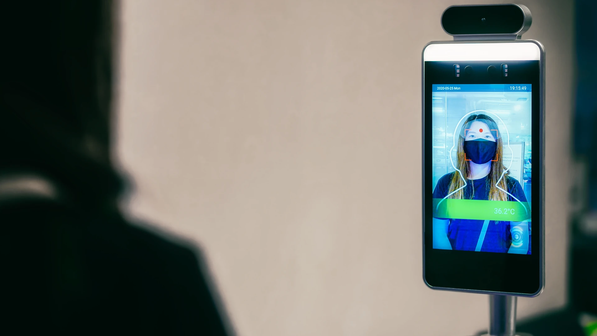 Contactless temperature-screening kiosk on a stand showing a masked person's face scan and temperature reading
