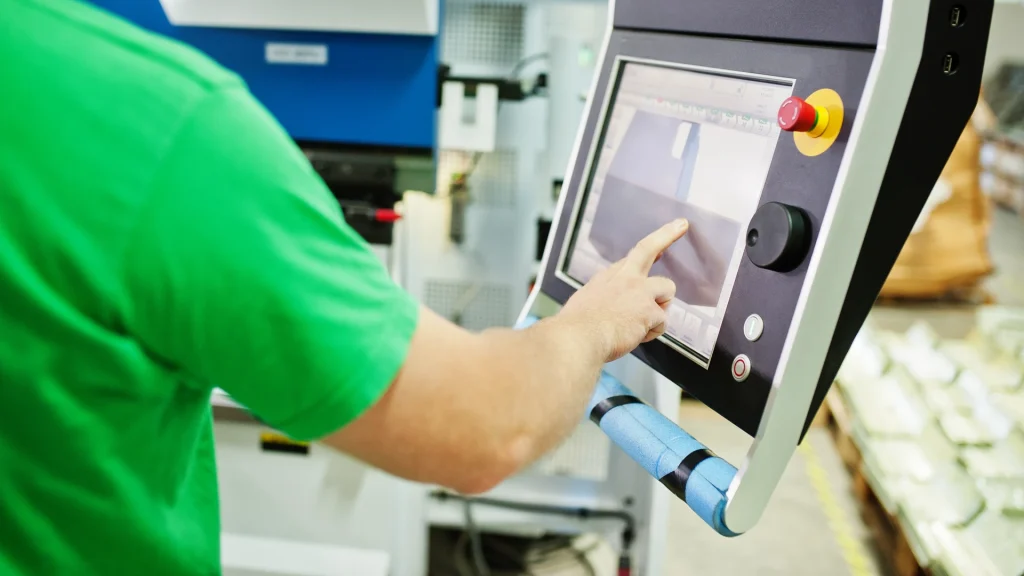 Machine operator in a green shirt touching a control-panel touch screen beside a red emergency-stop button and a black rotary knob.