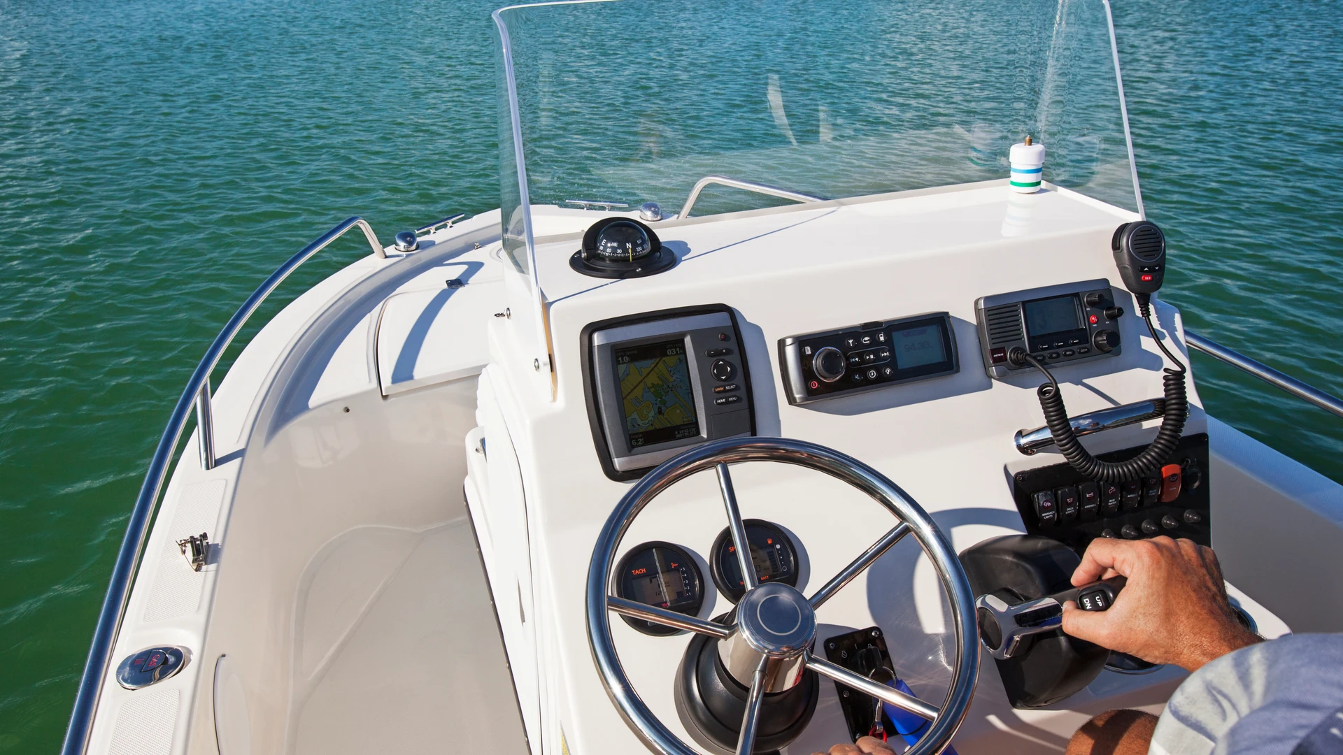 Hand on the throttle at a center-console boat helm with a chartplotter, stereo, VHF radio and engine gauges on blue-green water