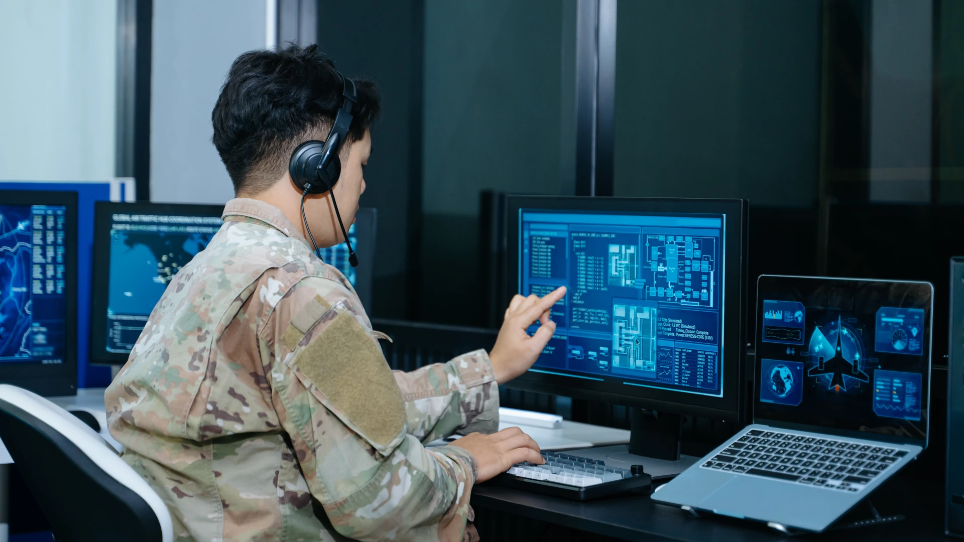 Operator in camouflage clothing wearing a headset and pointing at a monitor beside a laptop