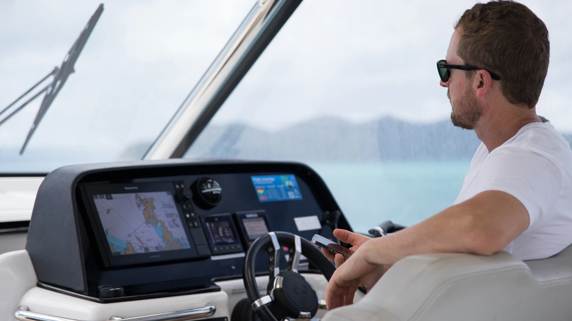 Boat driver in dark sunglasses seated at the helm beside a bright chartplotter showing a nautical chart in daylight