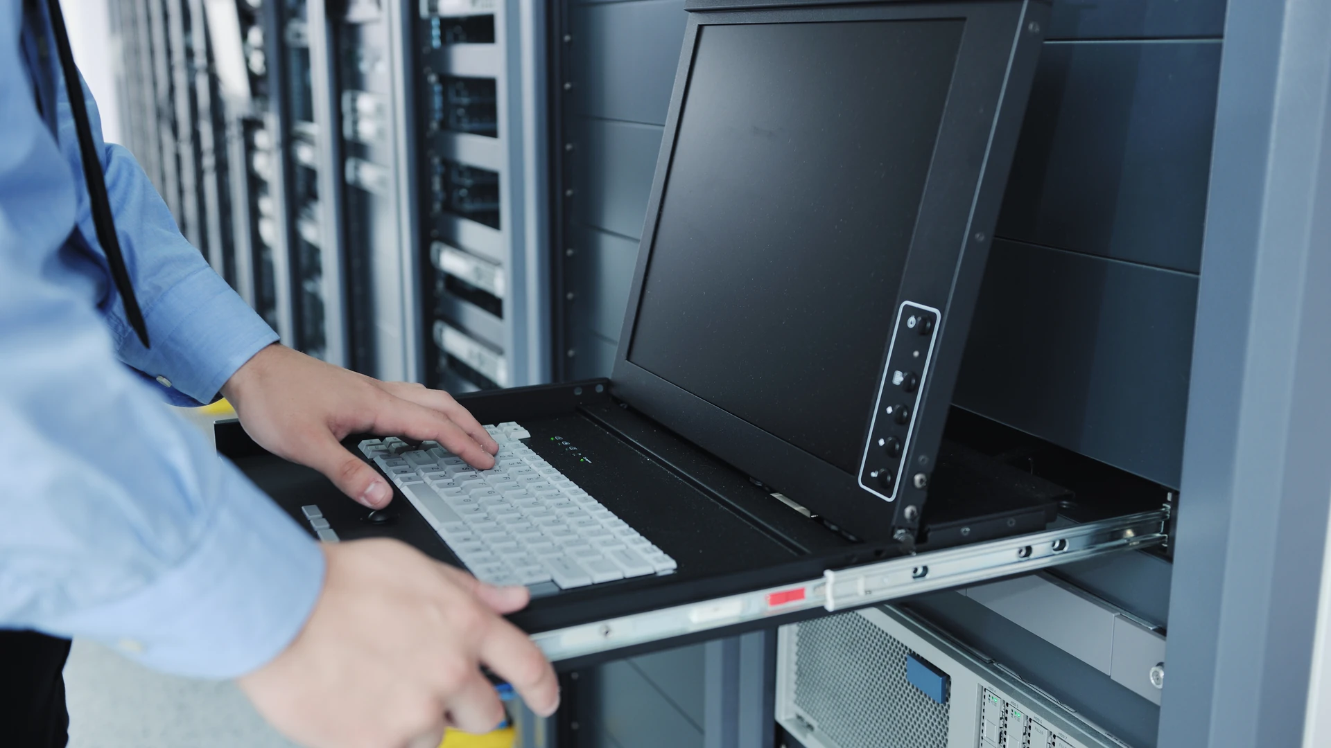 Hands on the keyboard of a console drawer pulled out of an equipment rack, with its flat screen raised and rows of server racks behind.
