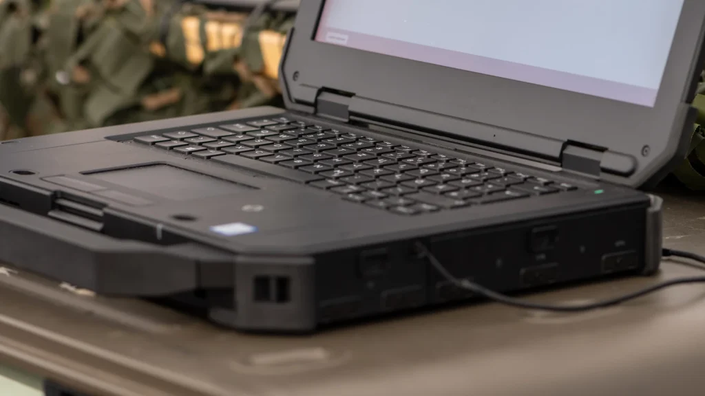Open rugged laptop with thick bumpers, hinge guards and a carry handle on an olive-green field case, camouflage netting behind.