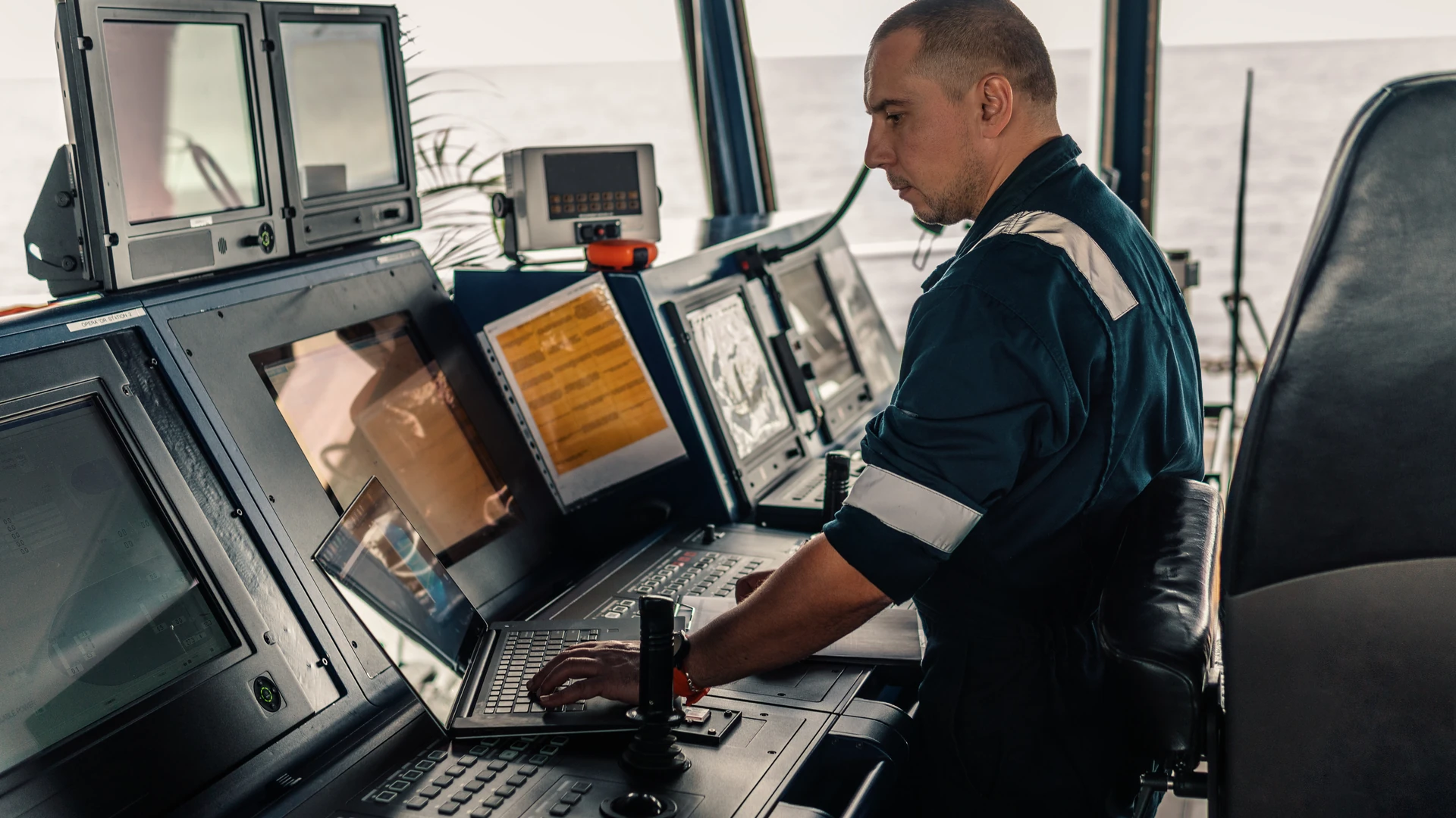 Officer at a ship's bridge console working on a laptop beside several displays, keypads and a joystick