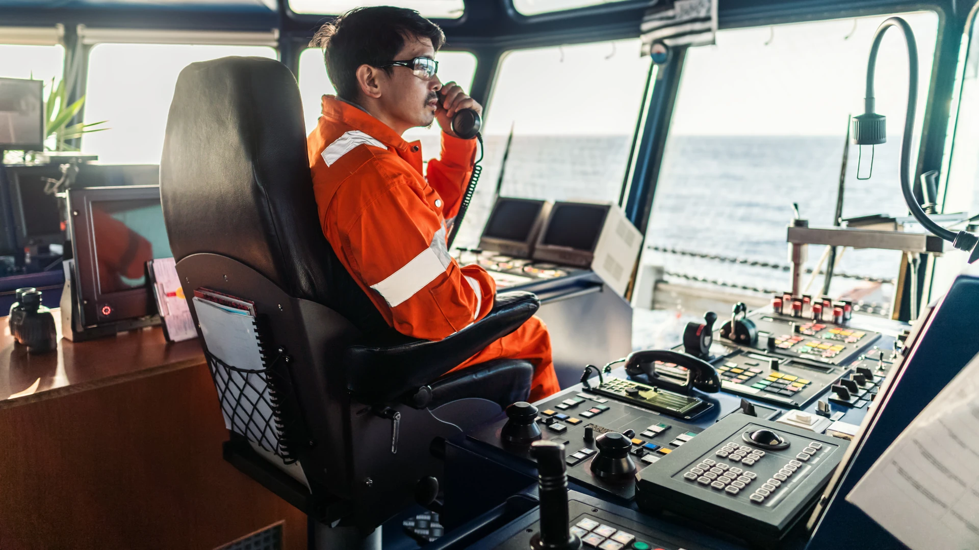 Deck officer on a ship's bridge speaking into a handset beside a console with keypads, a trackball and joysticks
