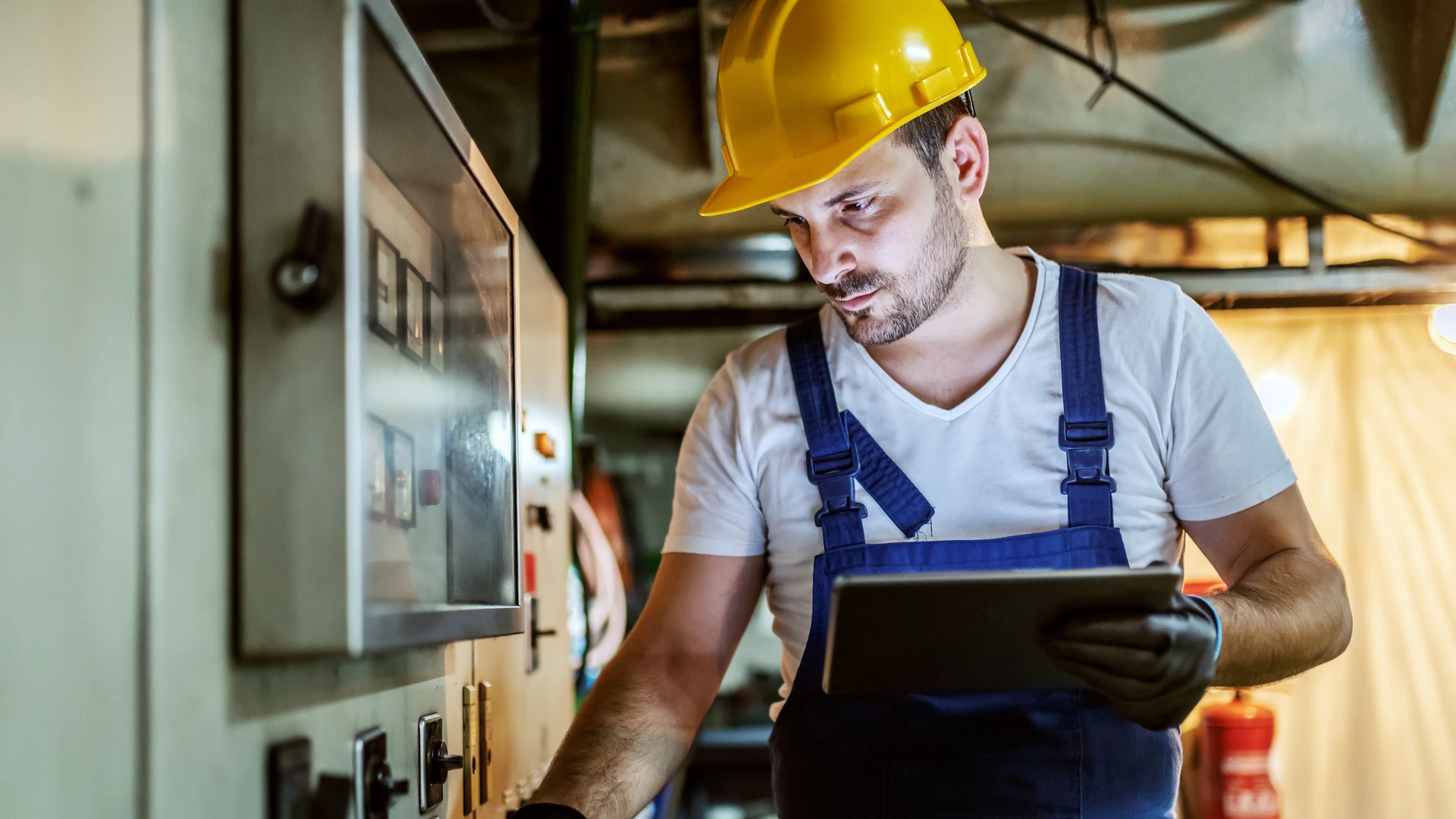 Worker in a yellow hard hat and blue overalls holding a tablet beside an industrial control panel